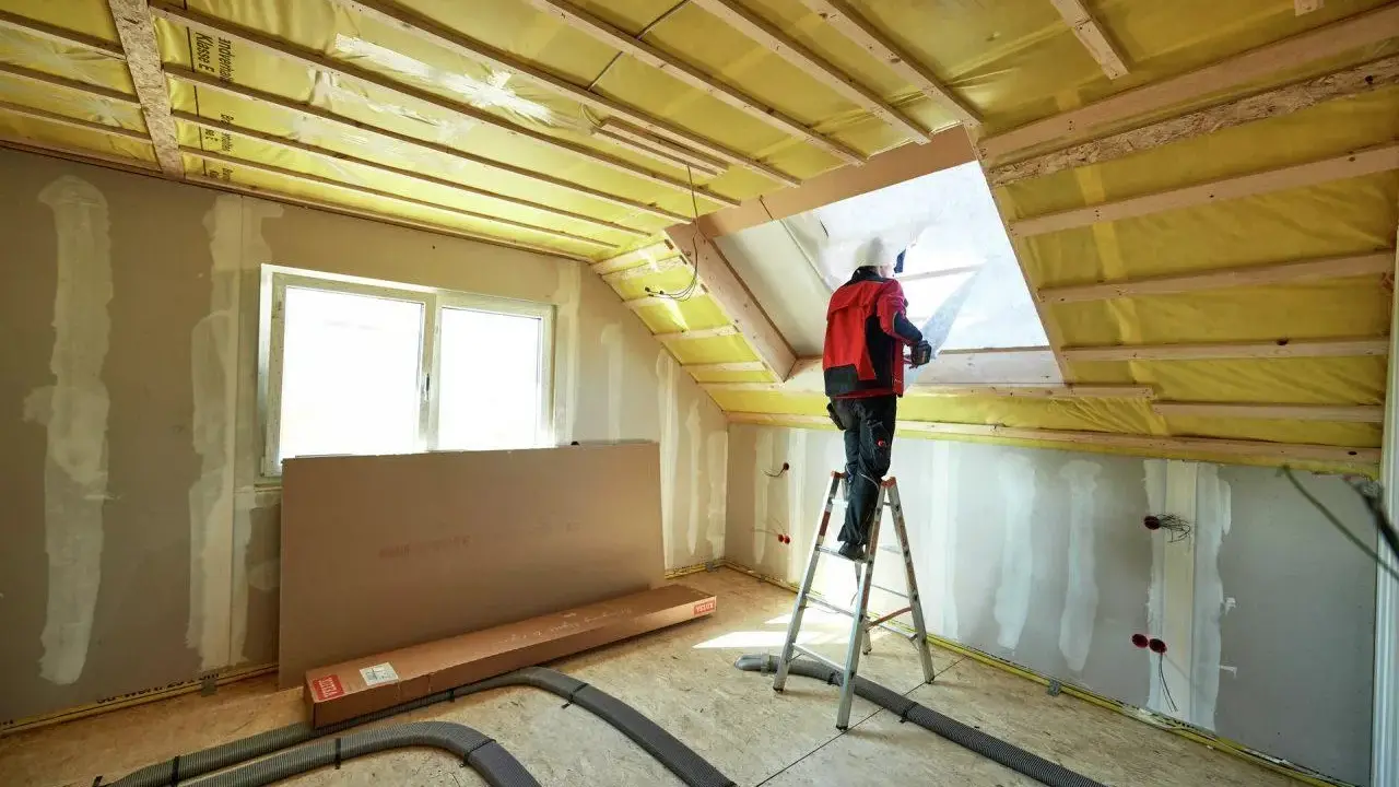 Lavori in corso per isolare sottotetto abitabile. Un operaio su scala installa materiale isolante giallo.