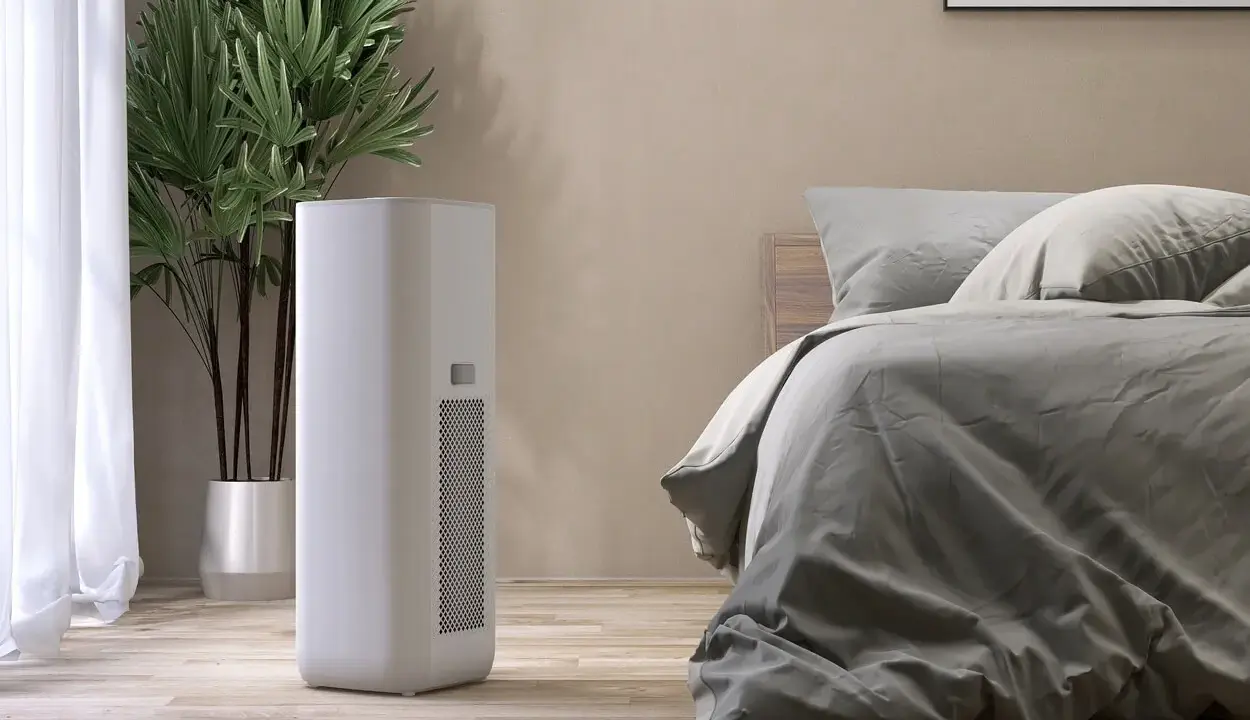 Un moderno deumidificatore o condizionatore bianco si trova accanto a un letto in una stanza accogliente, con una pianta verde sullo sfondo.