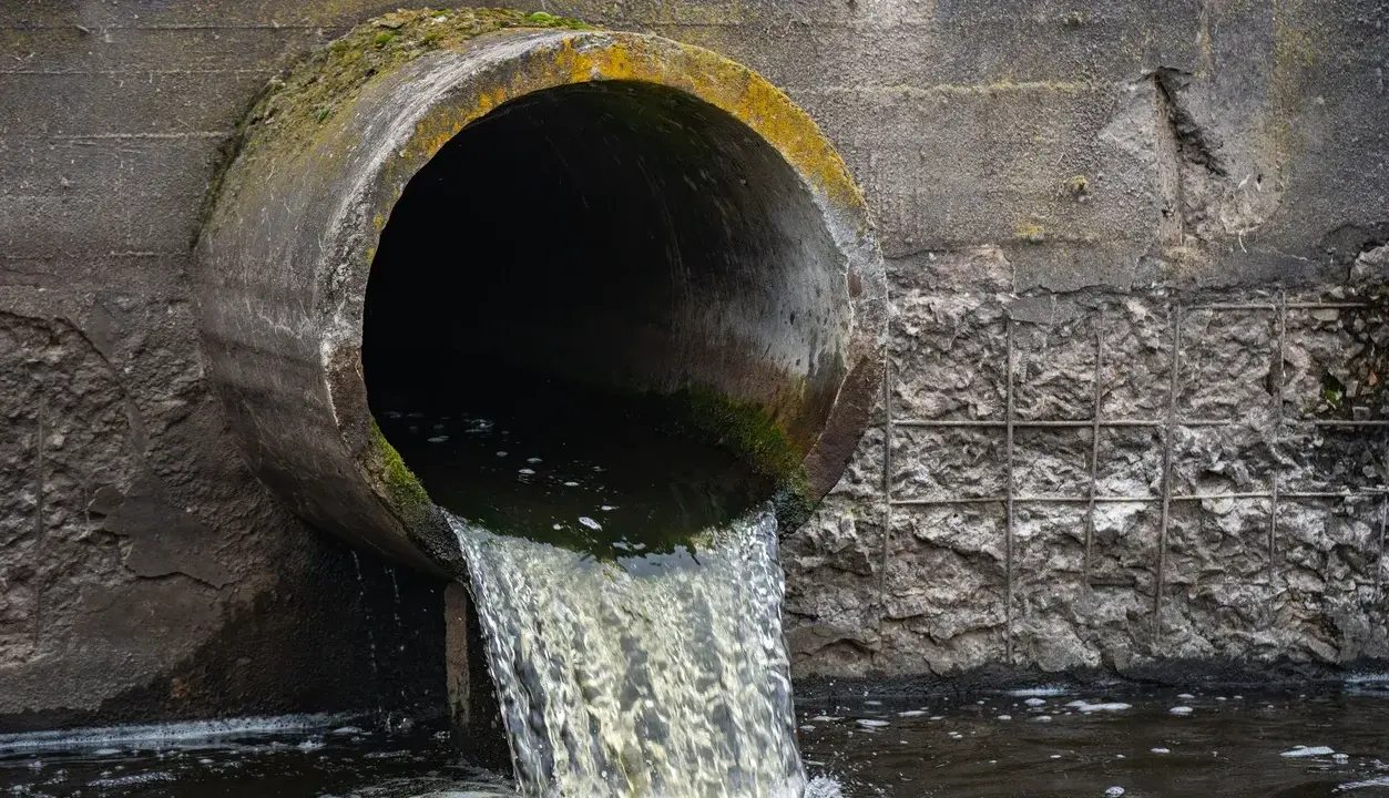 Un tubo di cemento scarica acque scure in un fiume. Le acque chiare del fiume si mescolano con quelle scure, creando un contrasto visivo.