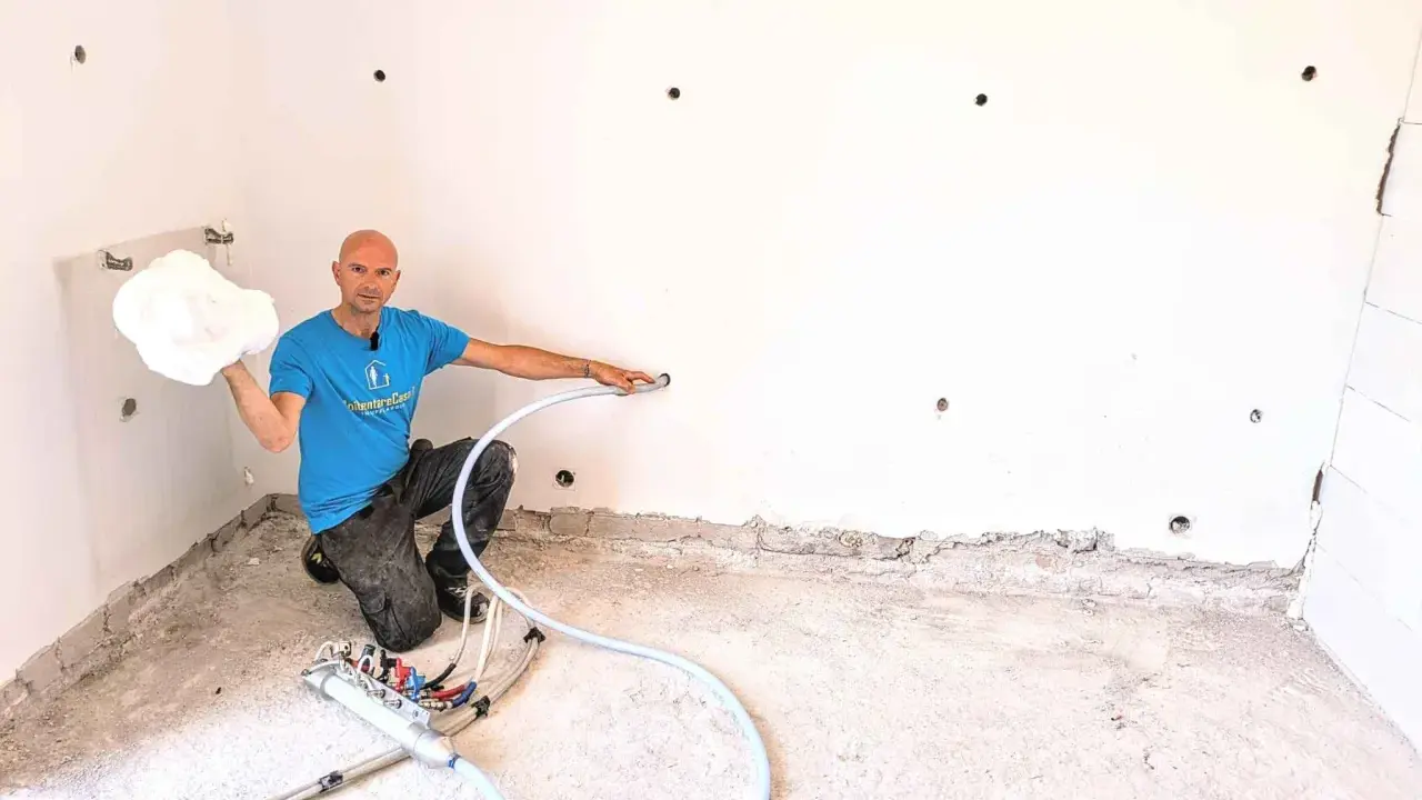 Man in blue shirt holding white foam, kneeling next to insulation equipment in a room with white walls and concrete floor.