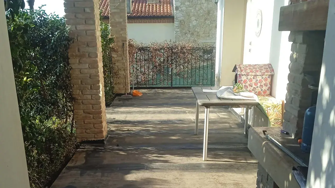 Terrazzo con pavimentazione scura, ideale per l'impermeabilizzazione. Tavolo, barbecue e vegetazione completano la scena.
