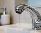 Un rubinetto cromato gocciola, un filo d'acqua che abbassa la pressione. Sullo sfondo, un dispenser di sapone e una pianta.