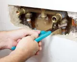 Mani con pinza lavorano su tubature in ottone. Si intravede una valvola di non ritorno prima o dopo la pompa, parte di un impianto idraulico.