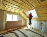 Lavori in corso per isolare sottotetto abitabile. Un operaio su scala installa materiale isolante giallo.