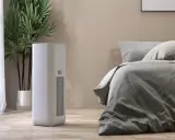 Un moderno deumidificatore o condizionatore bianco si trova accanto a un letto in una stanza accogliente, con una pianta verde sullo sfondo.