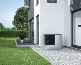 Unità esterna per riscaldamento con condizionatore, posizionata accanto a una casa moderna con giardino curato.