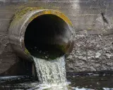 Un tubo di cemento scarica acque scure in un fiume. Le acque chiare del fiume si mescolano con quelle scure, creando un contrasto visivo.