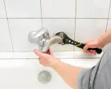 Mano con chiave inglese ripara un rubinetto che gocciola sopra una vasca da bagno bianca.