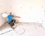 Man in blue shirt holding white foam, kneeling next to insulation equipment in a room with white walls and concrete floor.