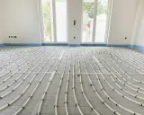 Installazione di riscaldamento a pavimento. Tubi bianchi disposti a serpentina su un foglio isolante, pronti per la posa del massetto. La temperatura dell'acqua sarà regolata per un comfort ottimale.