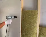 Mano con guanto bianco avvita cartongesso su struttura in legno, con isolamento in lana di roccia. Un esempio di isolamento acustico, che riduce il rumore a circa 75 decibel.