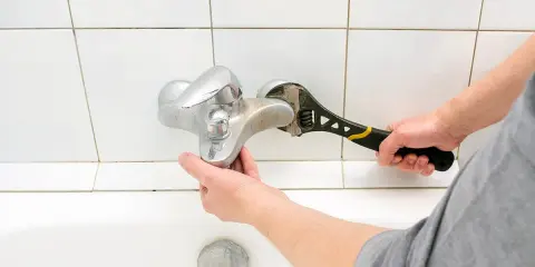 Mano con chiave inglese ripara un rubinetto che gocciola sopra una vasca da bagno bianca.