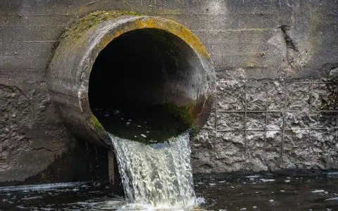 Un tubo di cemento scarica acque scure in un fiume. Le acque chiare del fiume si mescolano con quelle scure, creando un contrasto visivo.