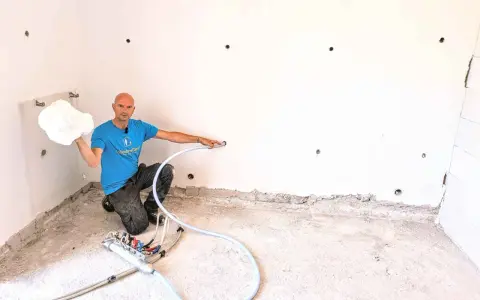 Man in blue shirt holding white foam, kneeling next to insulation equipment in a room with white walls and concrete floor.