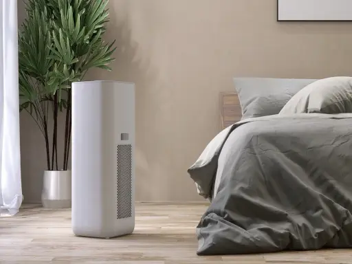 Un moderno deumidificatore o condizionatore bianco si trova accanto a un letto in una stanza accogliente, con una pianta verde sullo sfondo.