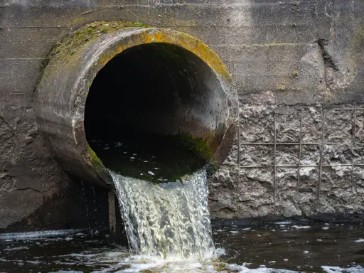 Un tubo di cemento scarica acque scure in un fiume. Le acque chiare del fiume si mescolano con quelle scure, creando un contrasto visivo.