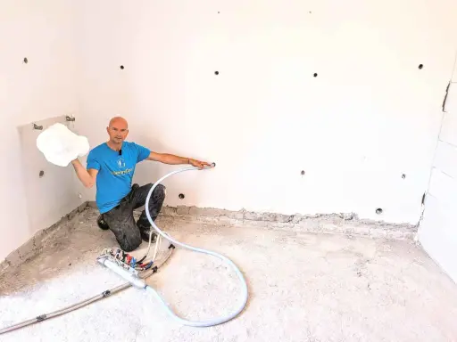 Man in blue shirt holding white foam, kneeling next to insulation equipment in a room with white walls and concrete floor.