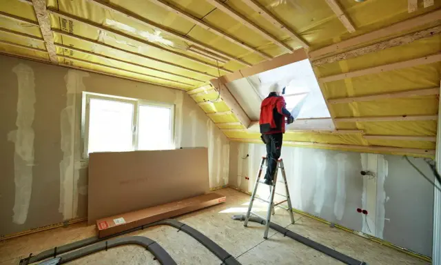 Lavori in corso per isolare sottotetto abitabile. Un operaio su scala installa materiale isolante giallo.