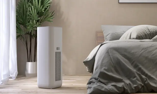 Un moderno deumidificatore o condizionatore bianco si trova accanto a un letto in una stanza accogliente, con una pianta verde sullo sfondo.