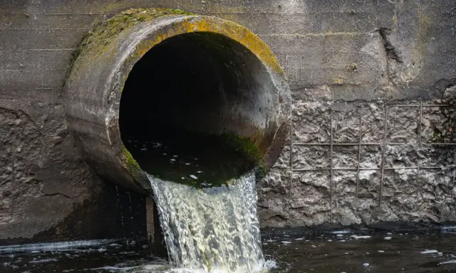 Un tubo di cemento scarica acque scure in un fiume. Le acque chiare del fiume si mescolano con quelle scure, creando un contrasto visivo.