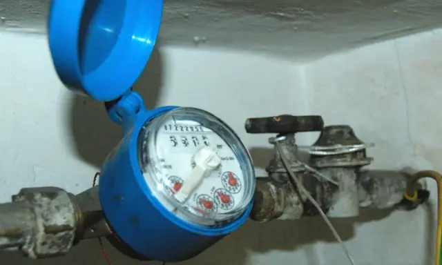 Contatore acqua blu con coperchio aperto, il quadrante è illeggibile per condensa. Tubi e rubinetto arrugginiti.
