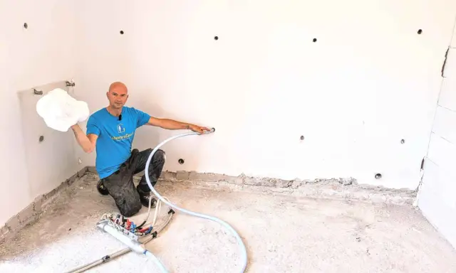 Man in blue shirt holding white foam, kneeling next to insulation equipment in a room with white walls and concrete floor.