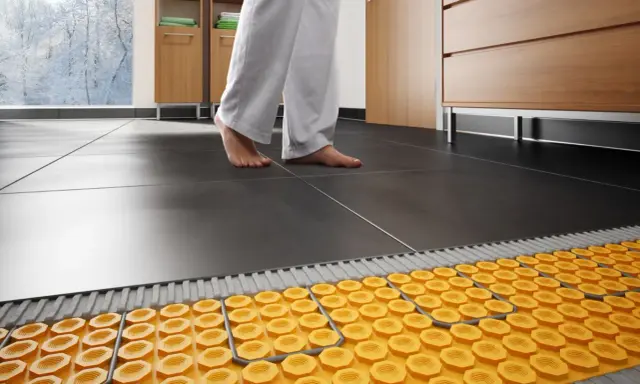 Piedi nudi su piastrelle scure, con un sistema di riscaldamento a pavimento visibile sotto. Addio termosifoni, benvenuta comodità!