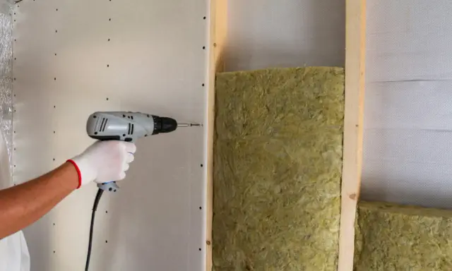 Mano con guanto bianco avvita cartongesso su struttura in legno, con isolamento in lana di roccia. Un esempio di isolamento acustico, che riduce il rumore a circa 75 decibel.