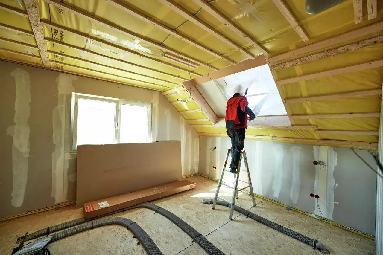 Operaio in piedi su una scala per isolare sottotetto con materiale isolante giallo.