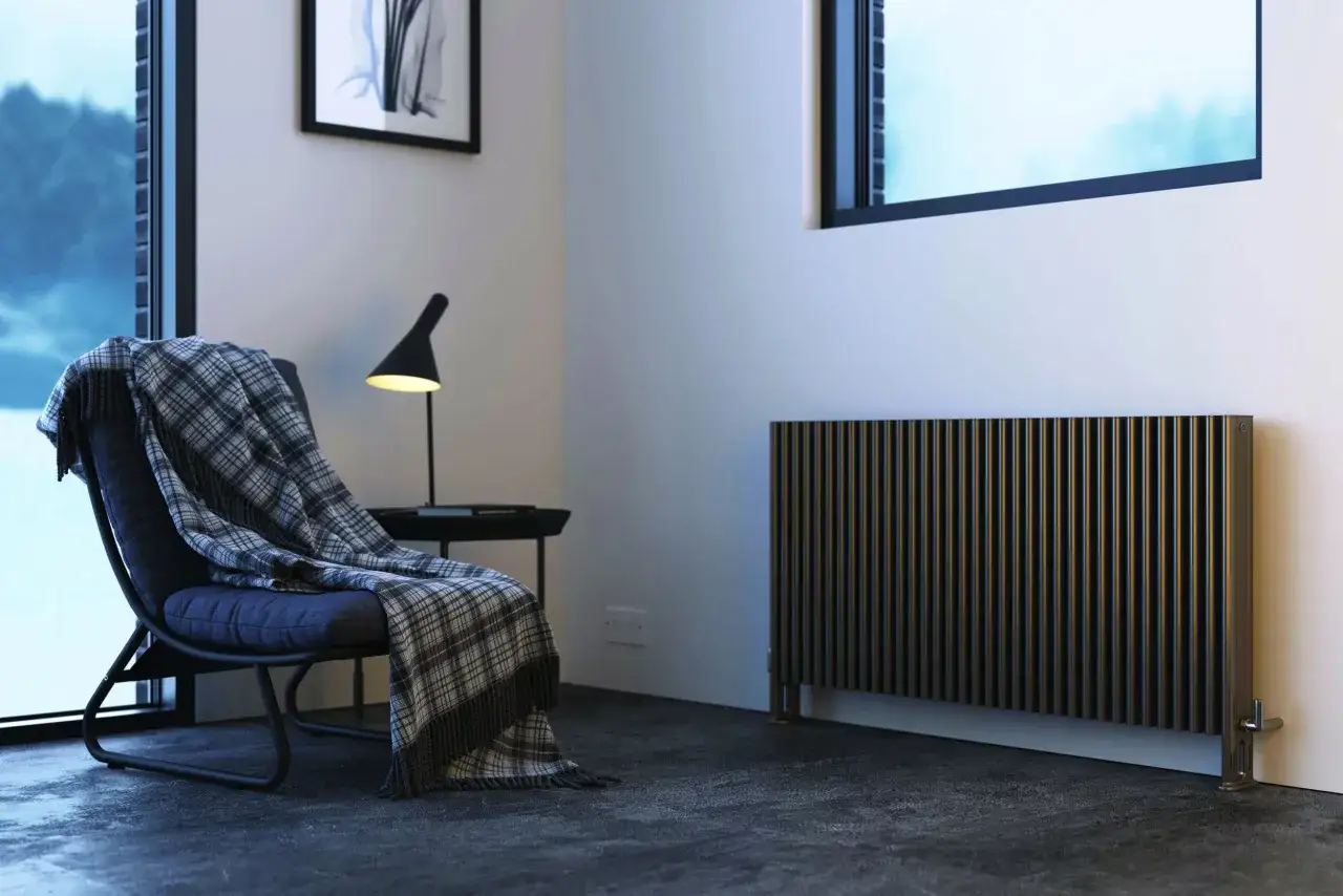 Un moderno radiatore nero, ideale per pompe di calore, riscalda un accogliente angolo lettura con poltrona e coperta.