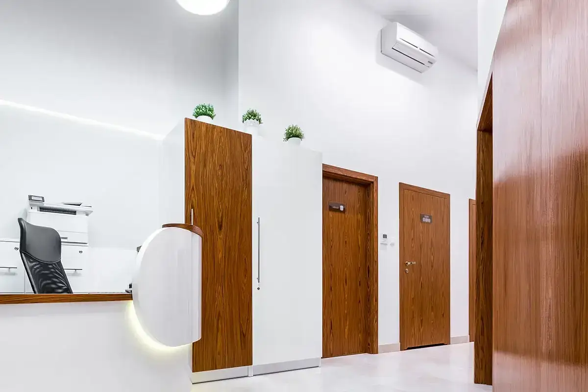 Un condizionatore d'aria a parete è installato in alto, pronto a rinfrescare due stanze. L'ambiente è moderno, con una reception e porte in legno.